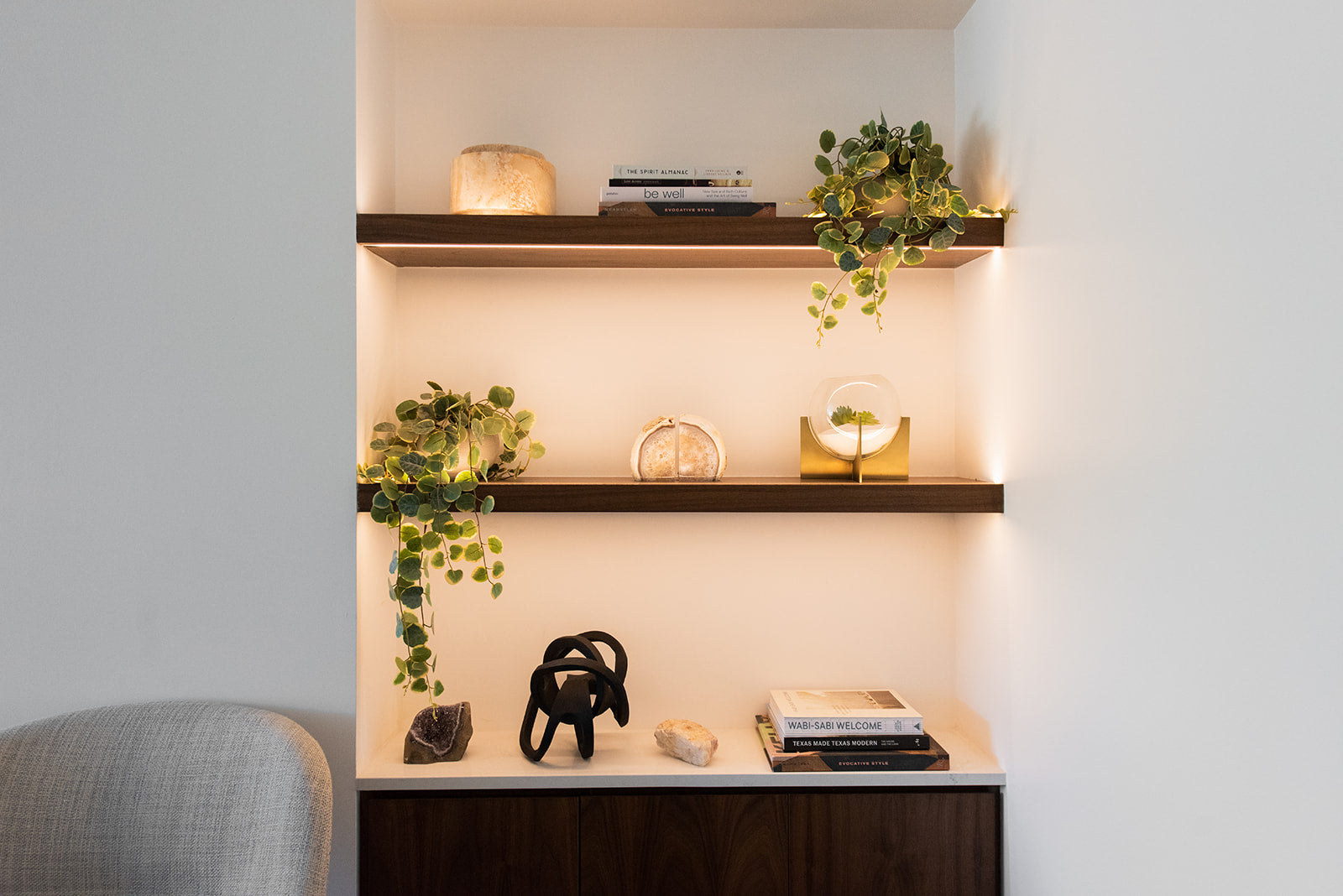 A built-in wall shelf unit with three dark wood floating shelves and warm under-shelf lighting, styled with books, small decor objects, and trailing green plants against a white wall.
