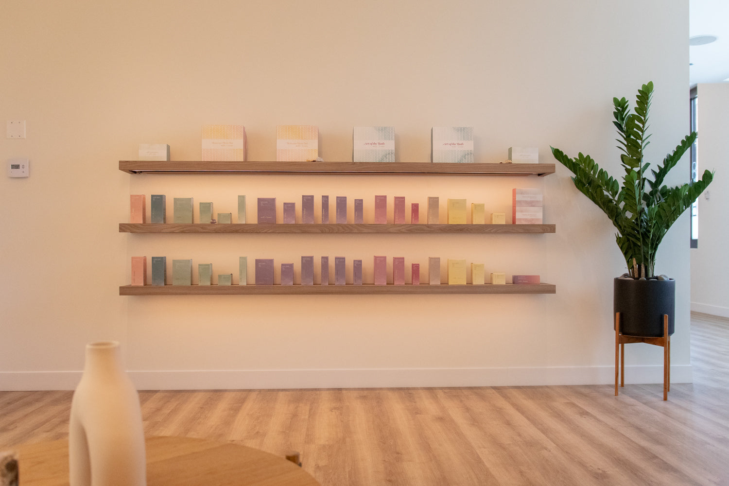 A styled interior scene showing three floating wooden shelves mounted on a cream wall, each holding rows of small pastel rectangular boxes or blocks. A potted green plant on a wooden stand stands to the right, and part of a beige table and vase are visible in the foreground.
