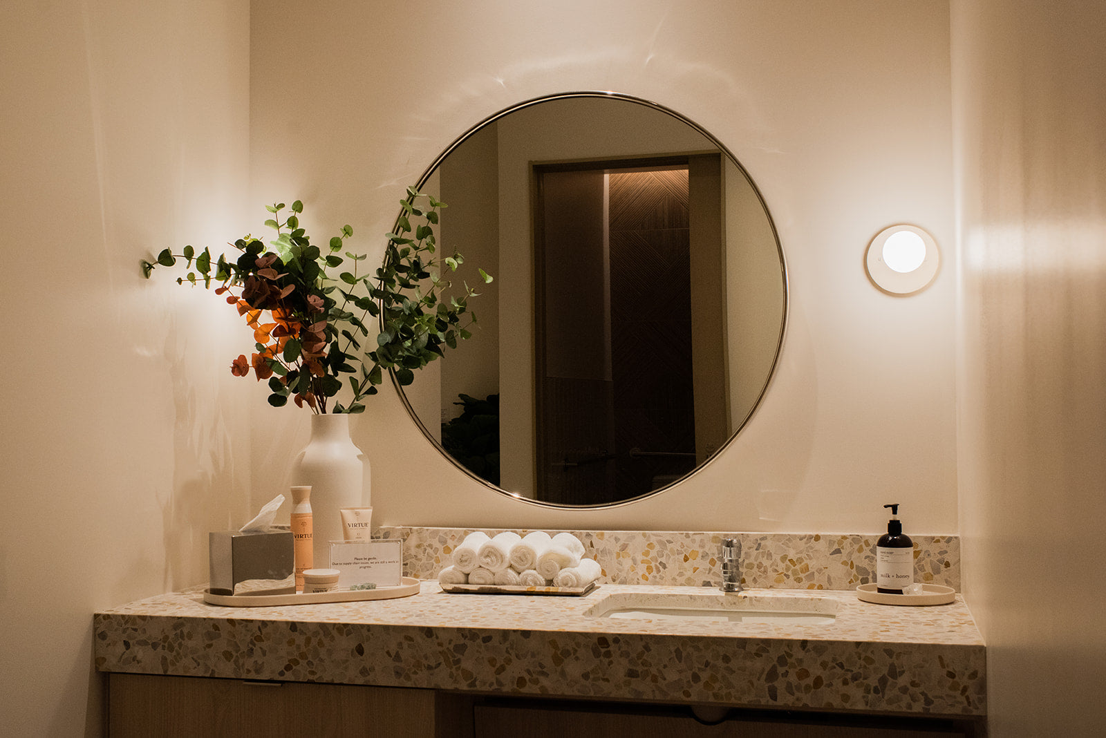 A warm, modern bathroom vanity setup featuring a large round wall mirror, a speckled stone countertop with a sink, a white vase of greenery and orange flowers, a tray of white bath towels, and a soap dispenser.