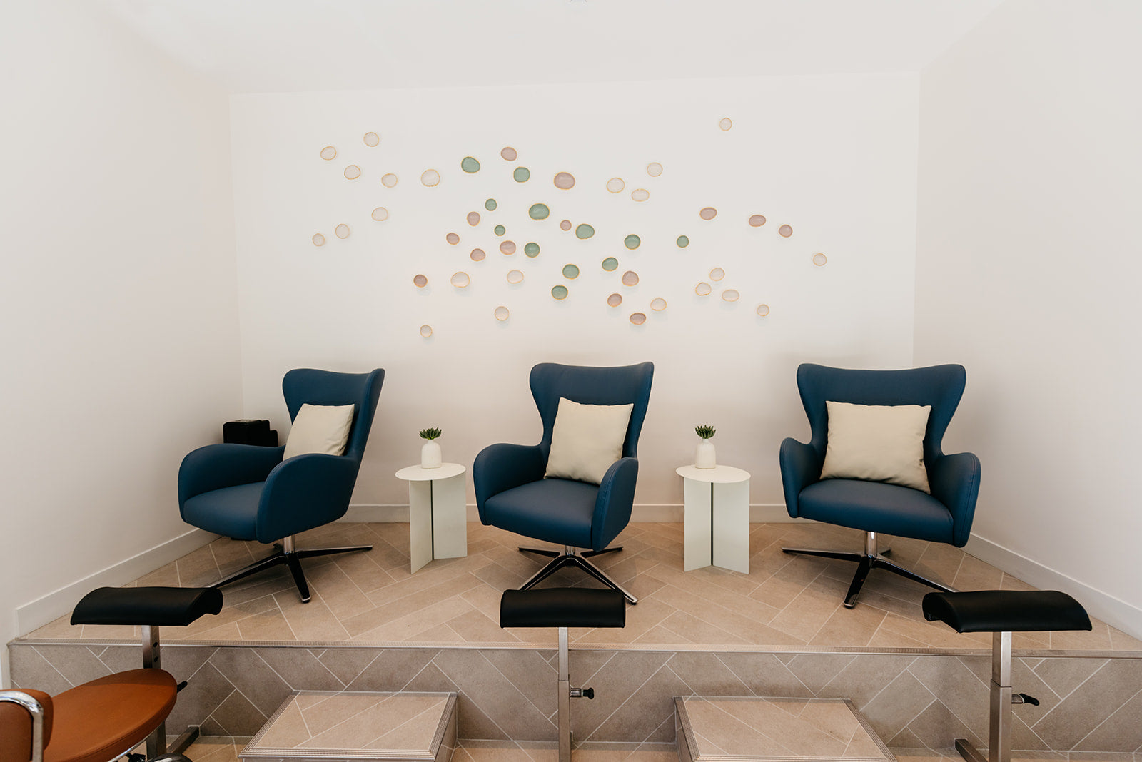 A staged interior room with three blue lounge chairs arranged on a raised platform, each with a cream pillow. Two small white side tables sit between the chairs, and a decorative cluster of circular wall decals is mounted on the white wall above.