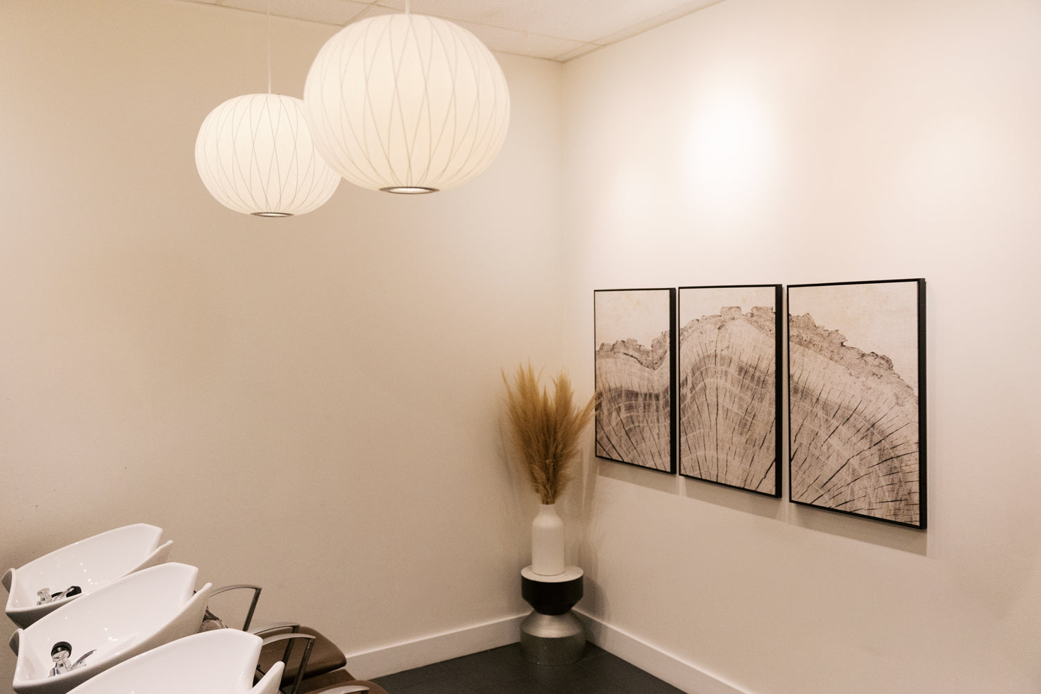A modern interior corner with two white spherical pendant lights hanging from the ceiling, cream walls, three framed abstract wood-art prints, a black-and-white pedestal, and a vase with dried grass. No people or animals are present.