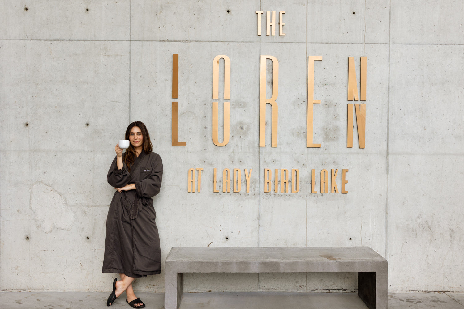 A woman in a dark brown robe stands next to a concrete bench in front of a concrete wall with large gold letters reading 'THE LOREM AT LADY BIRD LAKE'.