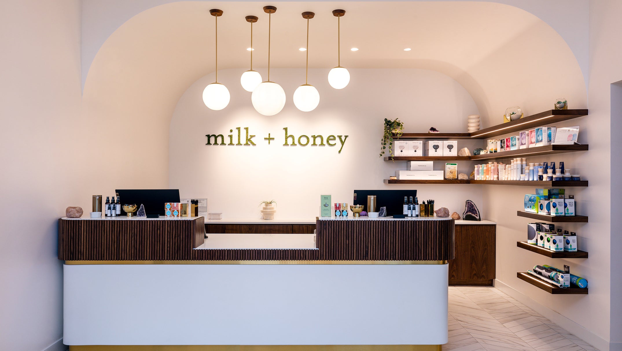 A modern retail counter and shelving inside a boutique with the words 'milk + honey' displayed on the back wall. The space features a white front counter with dark wood vertical slats, multiple spherical pendant lights, and open wall shelves stocked with products.