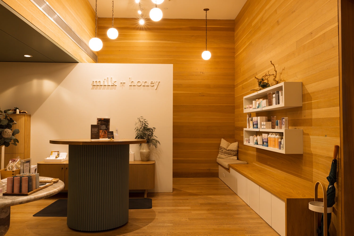 A warm, modern interior space with wood-paneled walls and floor, a round ribbed gray reception desk, white wall-mounted shelves with products, and a built-in bench with cushions against the right wall.