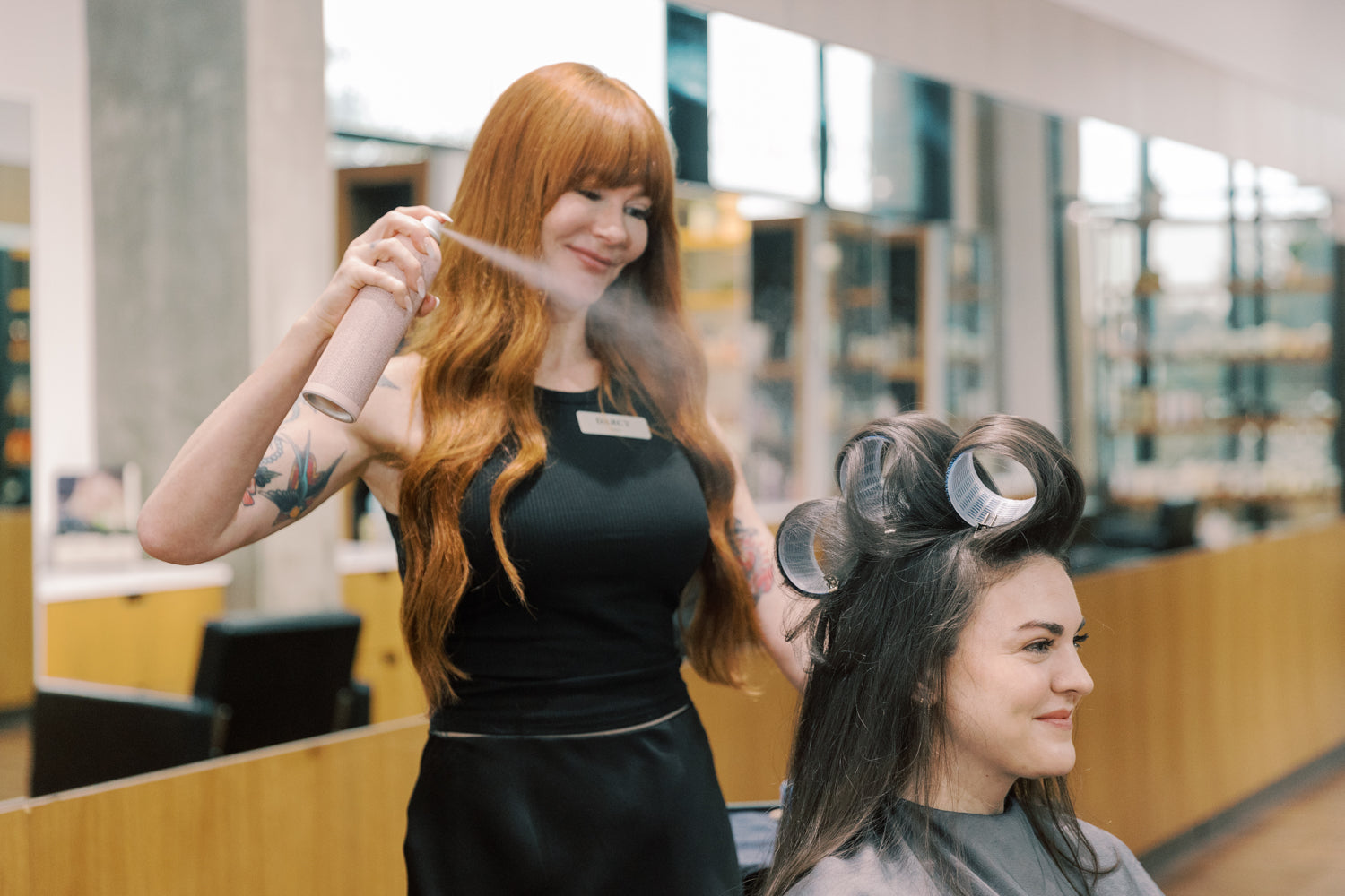 A woman with long auburn hair stands in a salon while another woman sits in the chair with curlers in her hair, receiving a hair spray treatment.