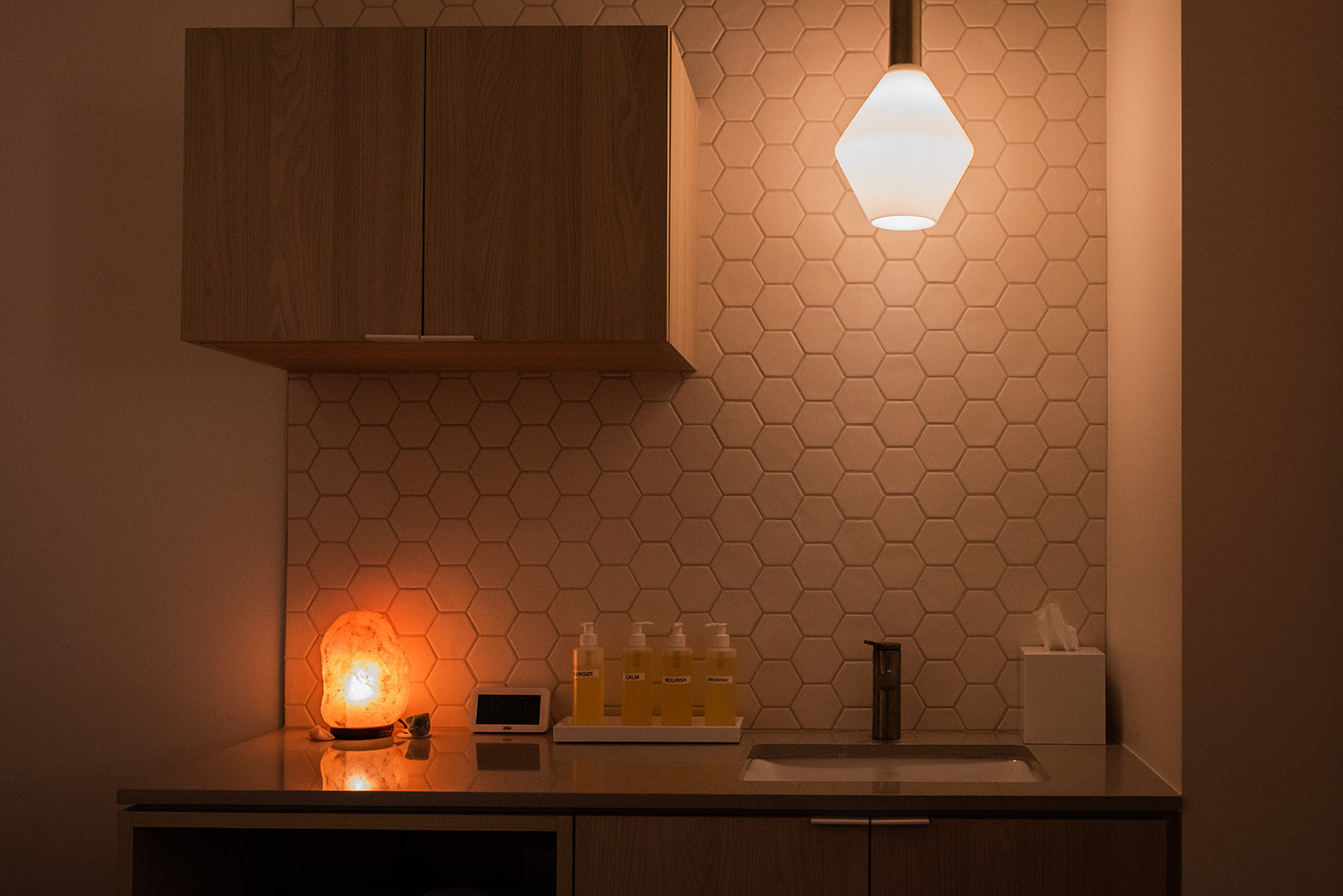 A modern bathroom vanity area with a hexagonal tiled backsplash, wooden cabinets, a countertop sink, and a glowing geometric pendant light above.