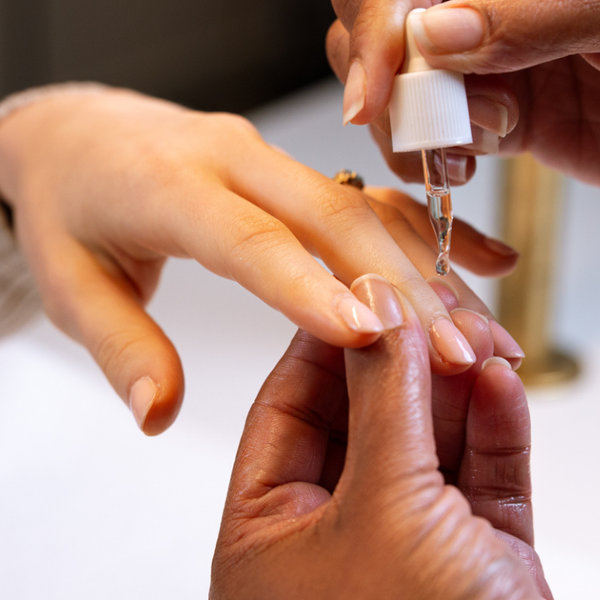 A close-up of a nail care dropper being used to apply oil to a fingernail, showing hands and a white dropper bottle.