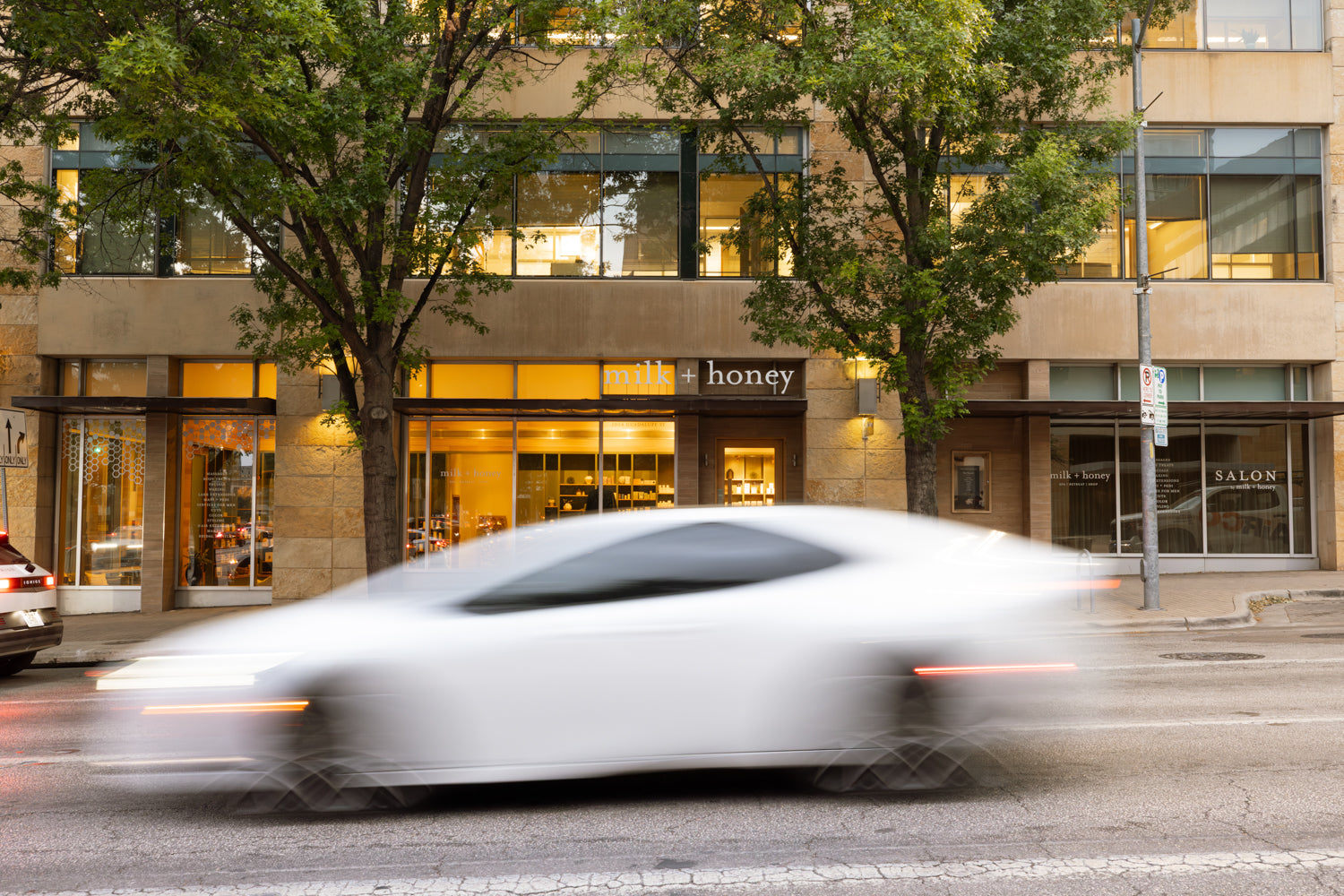 A blurred white sports car drives past a modern office building with storefronts, including a sign reading 'honey' and another reading 'SALON', with trees in front.