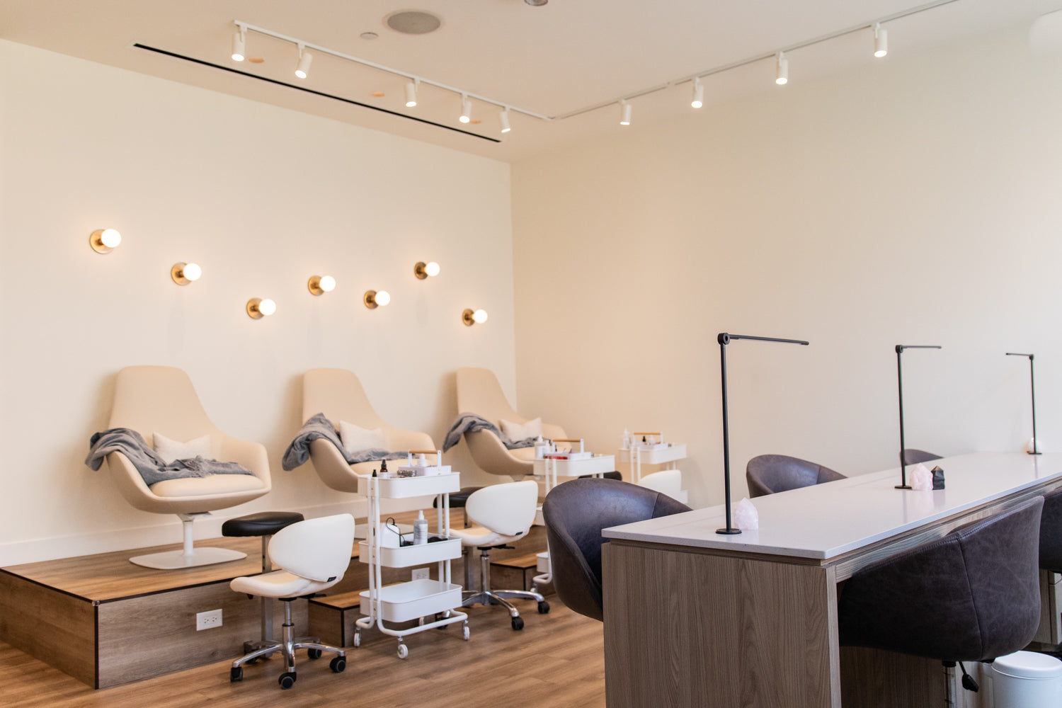A modern spa or salon interior with four beige reclining chairs along a wall, white treatment tables with black lamps, rolling stools, and warm wall lights on a light wood floor.