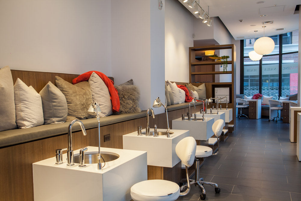 A modern spa or salon interior with multiple white sink stations, chrome faucets, white stools, a wood-and-cushion seating area with pillows, and large windows in the background.