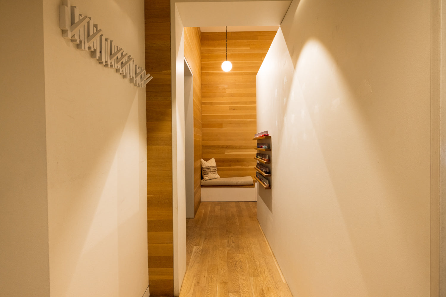 A modern interior hallway with warm wood flooring and walls, a built-in bench with pillows at the end, a hanging pendant light, and a decorative wall-mounted object on the left.