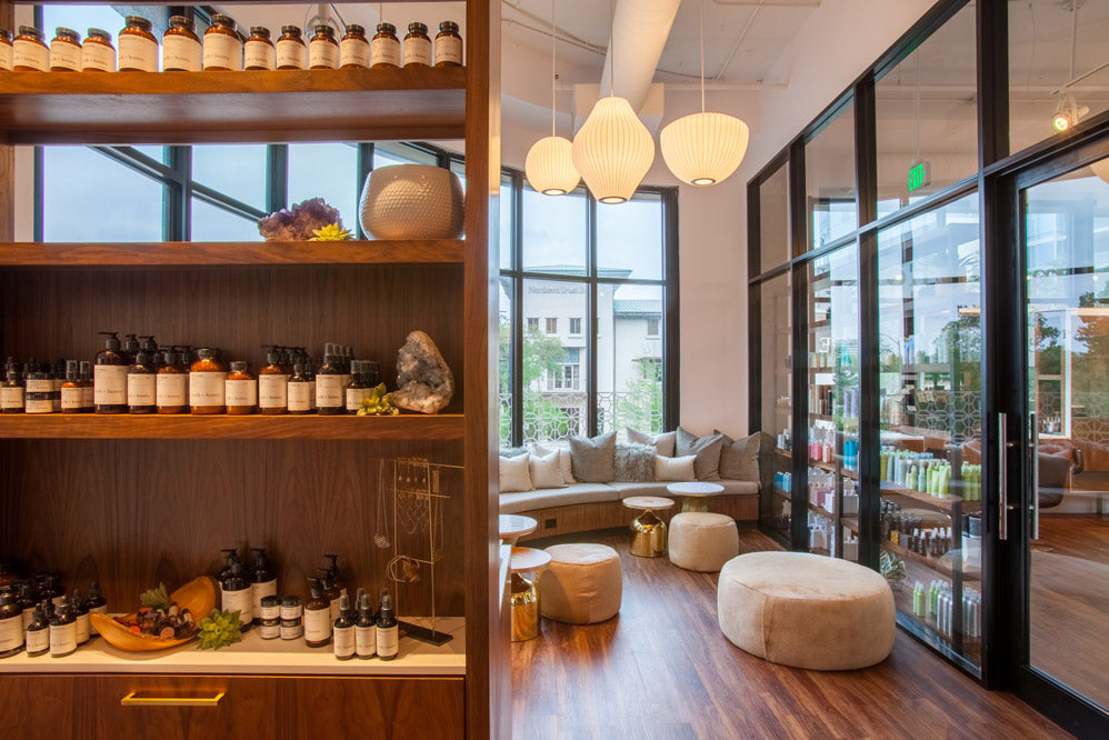 A modern wellness or spa retail interior with wooden shelving displaying many small bottles and jars, a curved seating area with cushions near large windows, pendant lights, and round ottomans on a wood floor.
