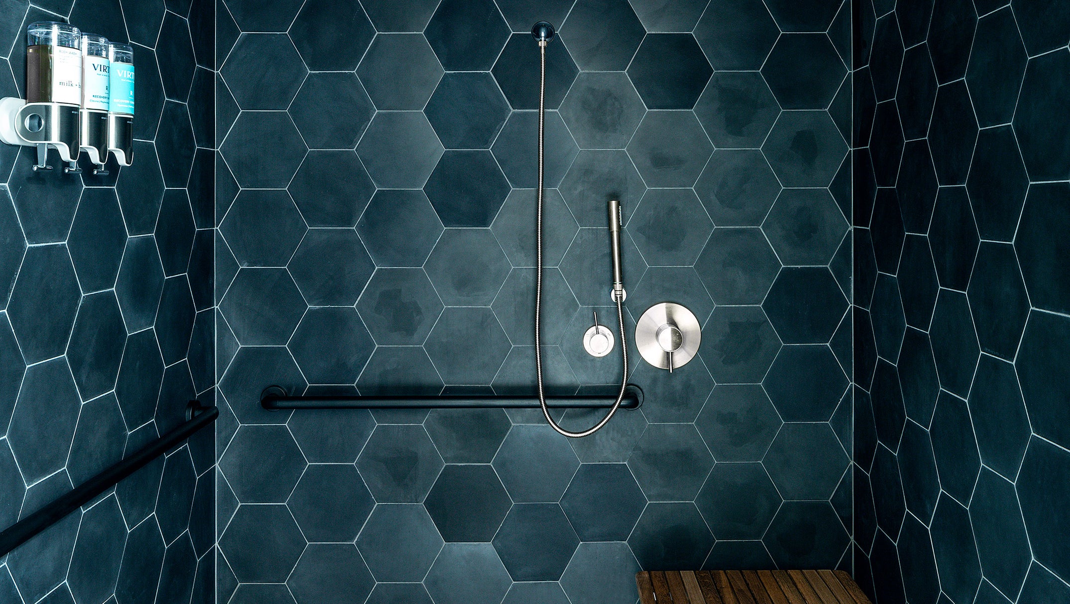 A modern walk-in shower stall with dark blue hexagonal wall tiles, a matching textured floor, a wooden bench mounted on the right wall, and a chrome shower fixture.
