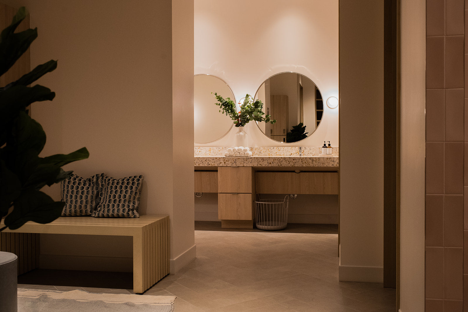 A modern bathroom interior with a double vanity, round mirrors, and warm ambient lighting, viewed from an adjacent hallway with a bench and plant in the foreground.