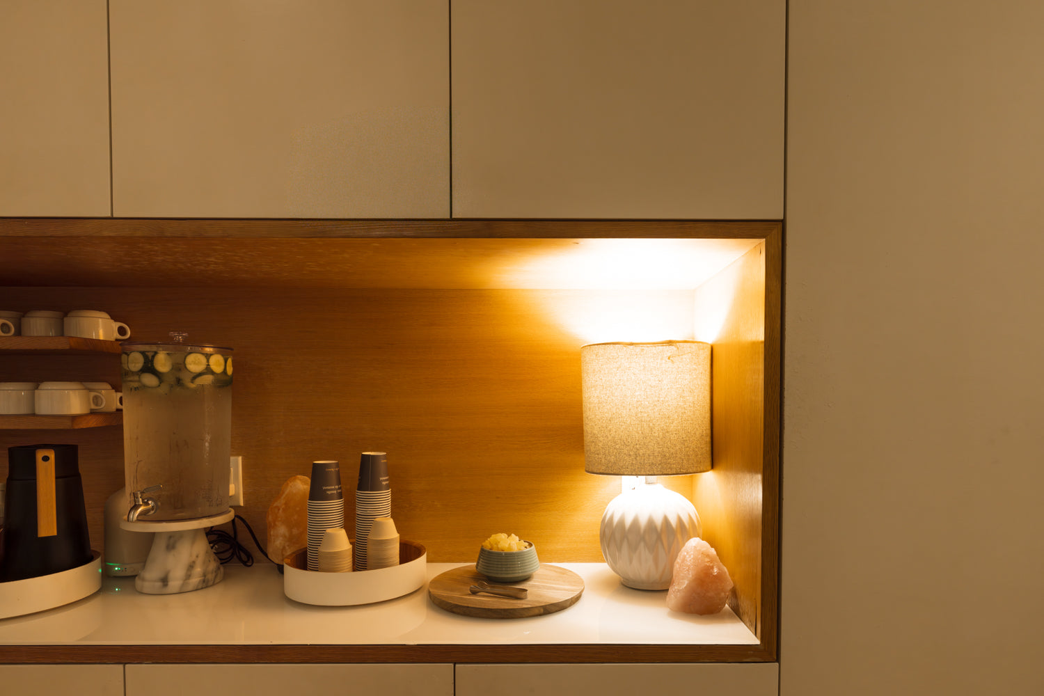 A warm, modern kitchen or bar counter with a built-in wooden shelf illuminated by a glowing lamp. The shelf holds a lit mug, a glass carafe, small bottles, a small plate with a yellow item, and a textured white table lamp with a pink crystal accent.