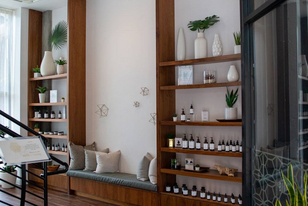 A styled interior scene showing wooden shelving units filled with small bottles, vases, and plants, alongside a cushioned bench with pillows in a bright room.