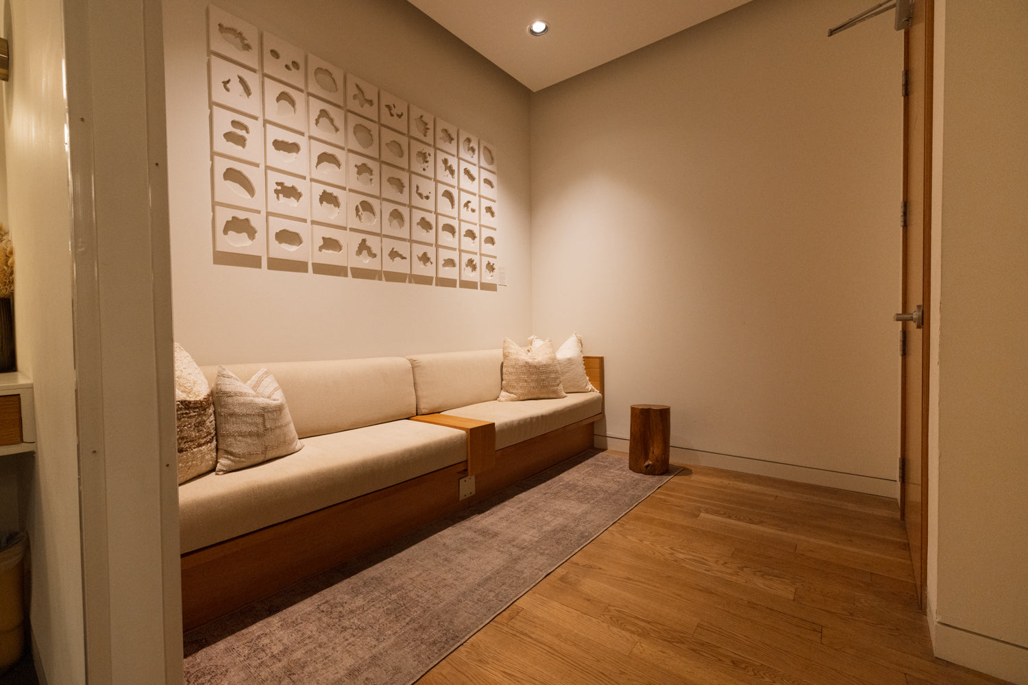 A modern low-profile beige sofa with a wooden base in a minimalist room, decorated with multiple square wall art prints and a gray rug on a wood floor.