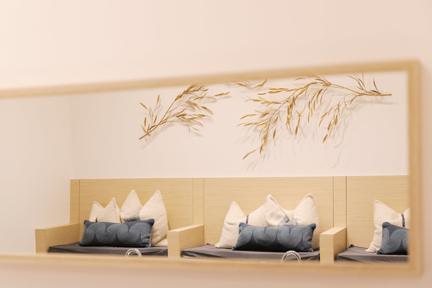 A minimalist interior scene showing a light wood bench with white and blue-gray pillows against a cream wall, with a large rectangular mirror above reflecting the scene and gold-toned dried botanical branches mounted on the wall.