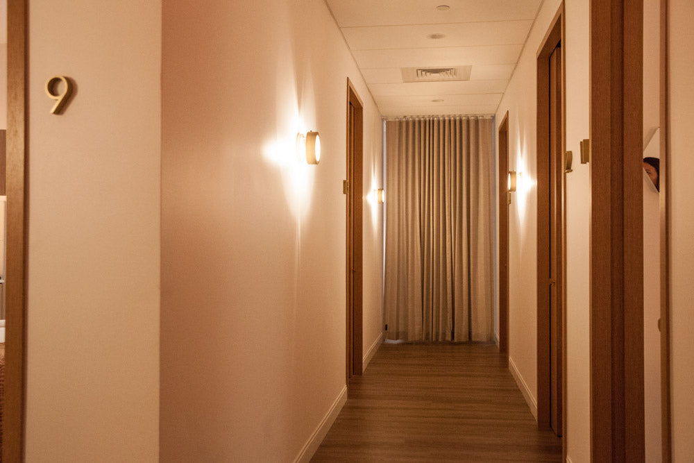 A warm, softly lit hotel hallway with cream-colored walls, wooden doors, a wood floor, wall-mounted sconces, and a beige curtain at the end of the corridor.
