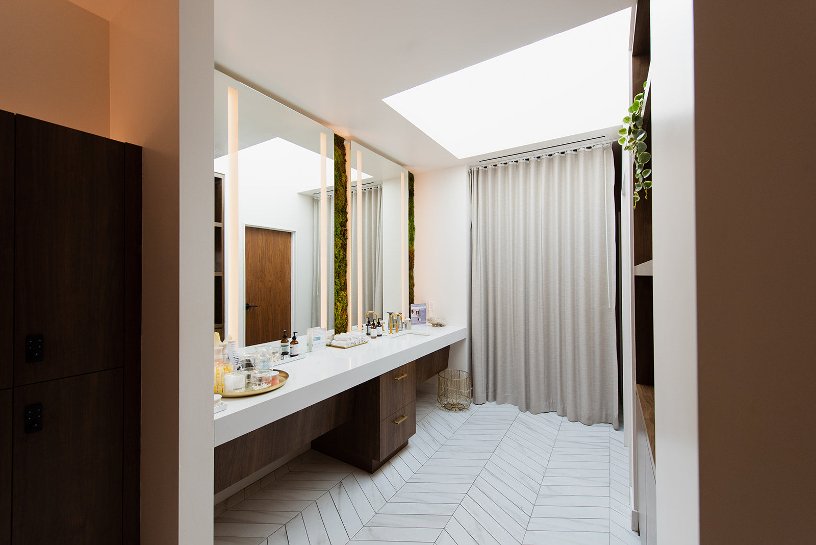 A modern bathroom interior with a long white double vanity, large mirrors, light wood cabinetry, white herringbone tile flooring, and a white pleated shower curtain under a skylight.