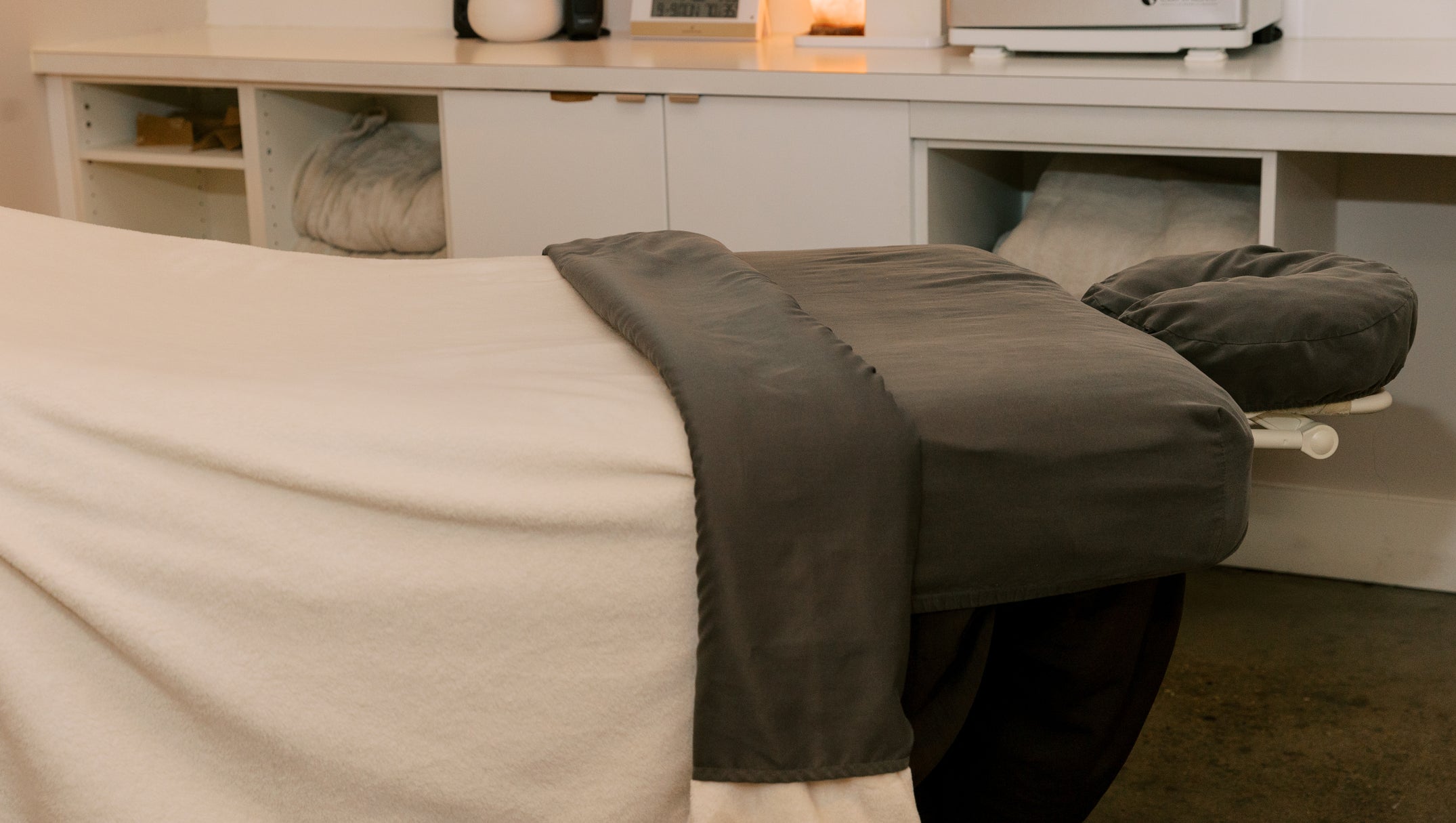 A massage table in a spa setting, covered with a white sheet and a dark gray towel, with a matching headrest. In the background, a white treatment counter holds a digital clock, a lit candle, and spa equipment.