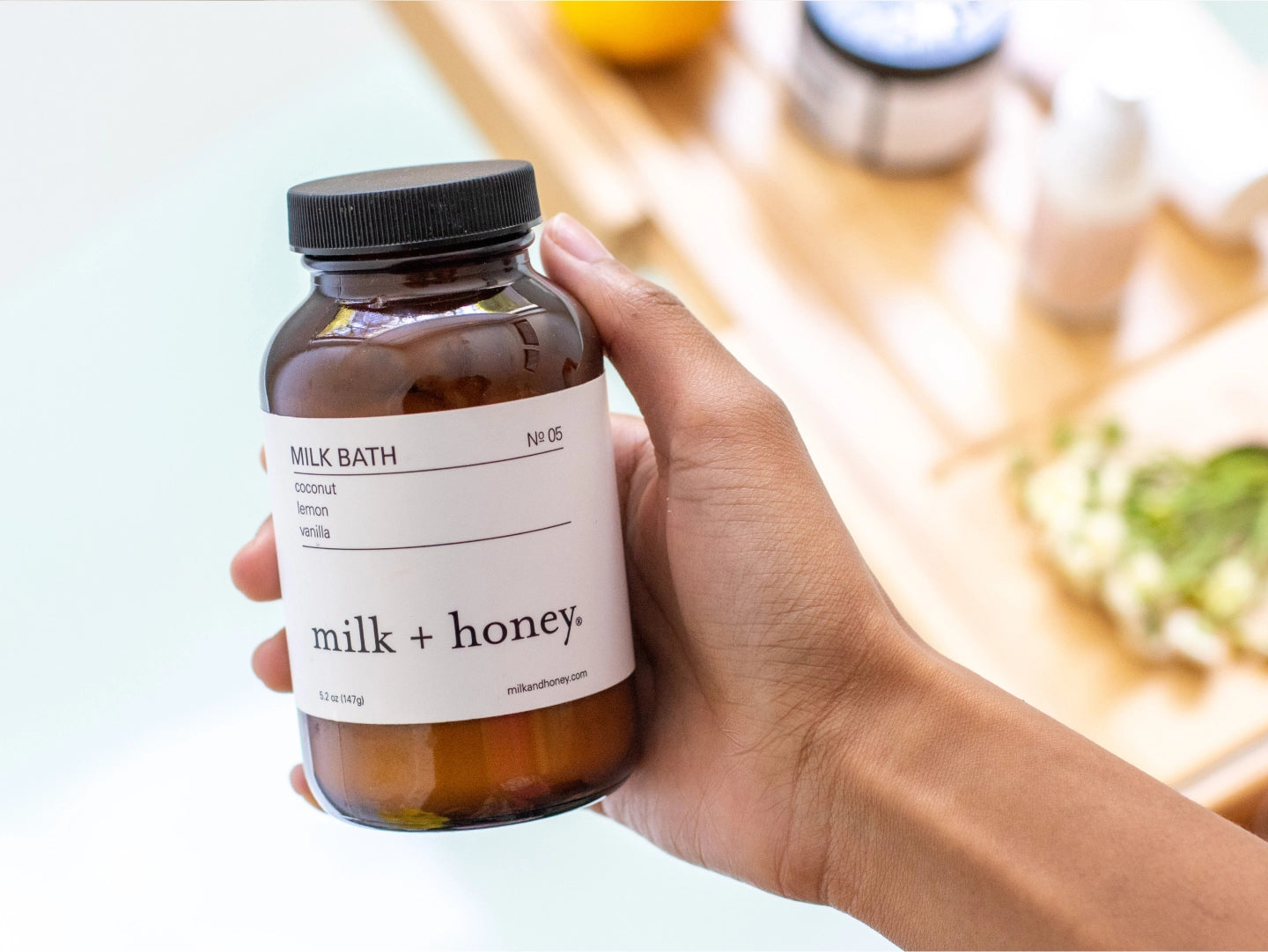 Hand holding a bottle of 'Milk + Honey' milk bath with a blurred background