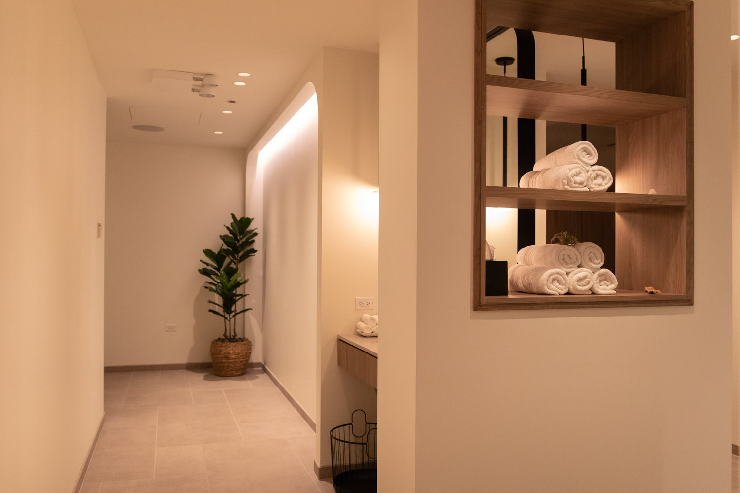 A warm, modern spa or bathroom interior with beige walls, a potted green plant in a hallway, and a built-in wooden shelf on the right holding neatly rolled white towels under warm lighting.