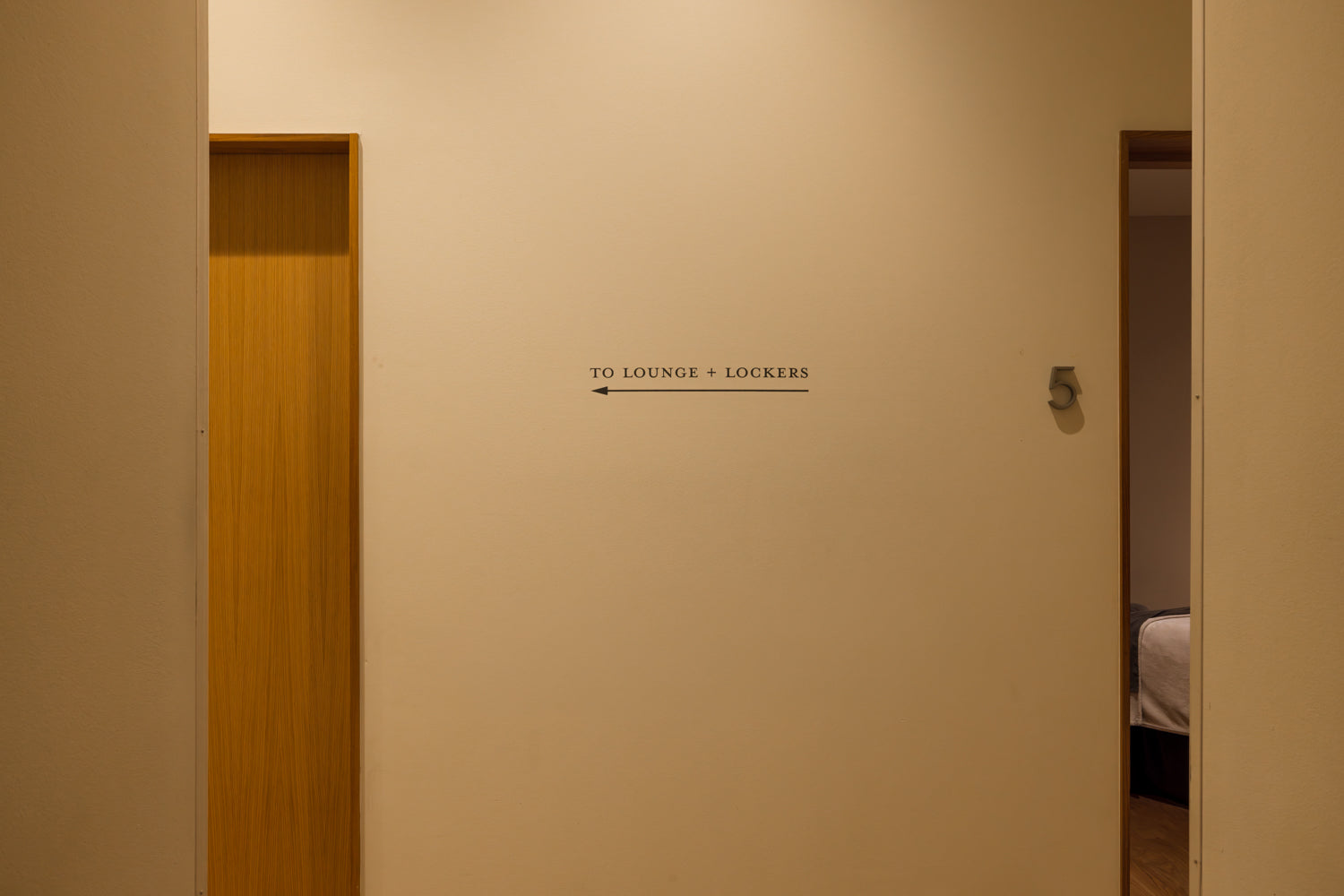 A beige interior hallway wall with a small black sign reading 'TO LOUNGE + LOCKERS' and an arrow pointing left, with a wooden door on the left and an open doorway on the right showing a bed.
